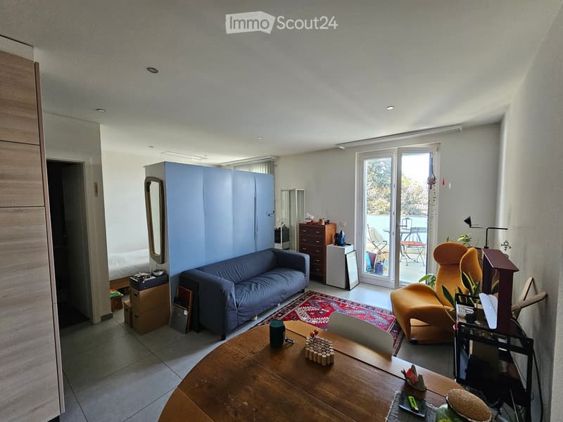 A modern living room with a blue sofa, orange armchair, wooden coffee table, and a view to a balcony. The room features a large sliding glass door leading to the balcony, a patterned rug, and a small cabinet with a mirror.