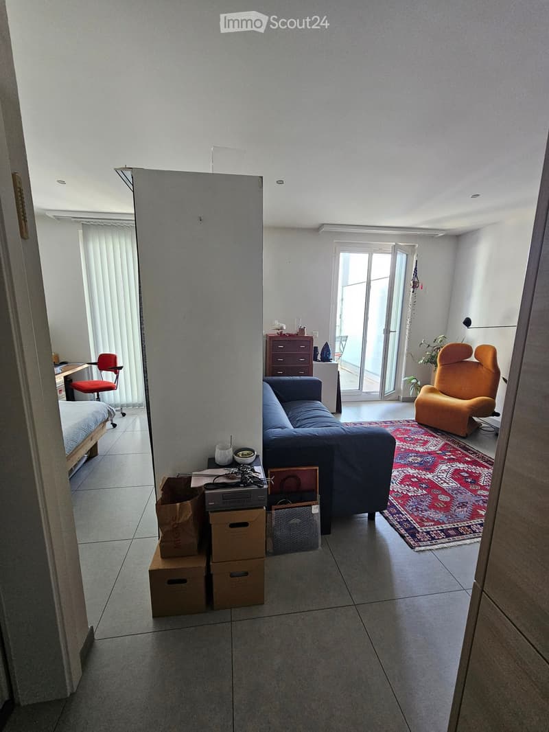 A modern living room with a blue sofa, orange armchair, small table with a bowl, a drawer unit, a bed visible in the background, and a large window with blinds. The room features a patterned rug and several boxes stacked near the entrance.