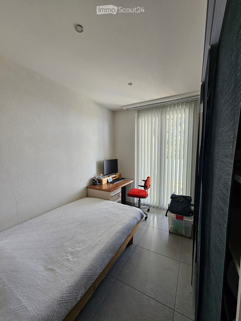 A small, furnished bedroom with a single bed, a wooden desk with a laptop and screen, a red office chair, and a backpack on the floor near the window with vertical blinds.