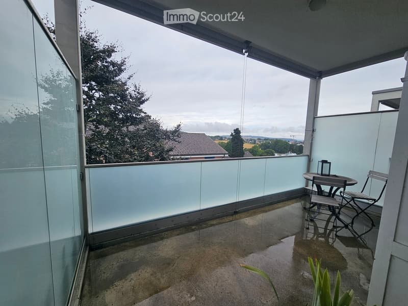 Balcony with glass railing, table, chairs, lantern, and a view of the surrounding area.