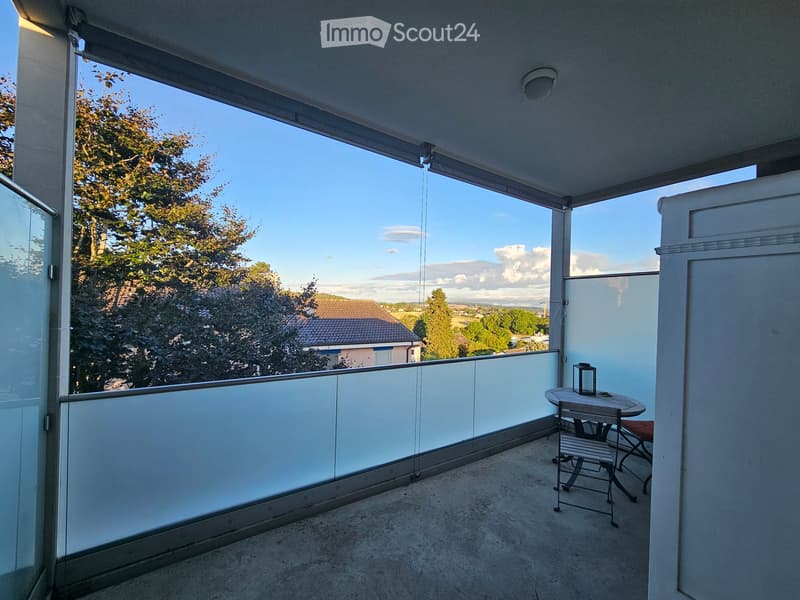 Balcony with glass railing, table, and chair, overlooking a scenic view of trees and a house.