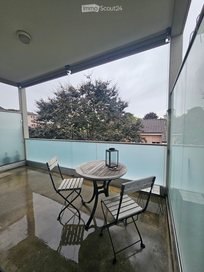 Balcony with a small round wooden table, two folding chairs, and a lantern, overlooking a residential area with trees and houses.
