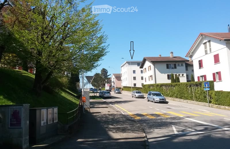 Residential street with multi-story buildings, parked cars, and greenery on both sides.