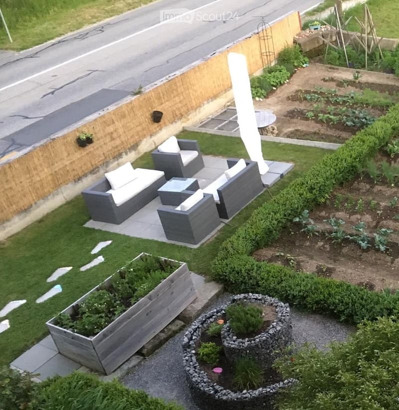 Garden with a seating area featuring a grey sofa, a table, and a chair, surrounded by plants and a wooden fence.