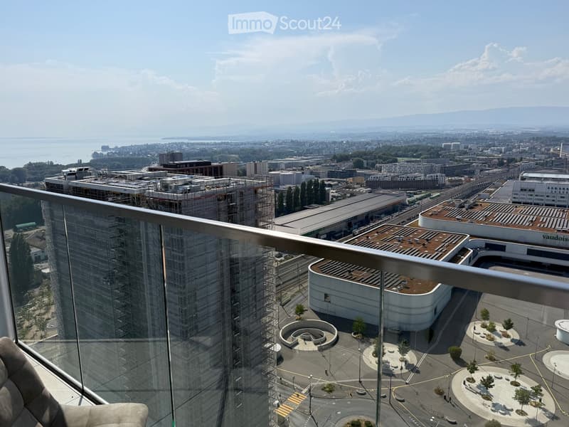 Panoramic city view from a high-rise balcony, showing urban landscape, water body, and distant mountains.