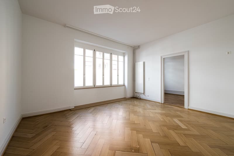 Empty room with wooden herringbone flooring, white walls, large window with multiple panes, radiator, and a doorway leading to another room.
