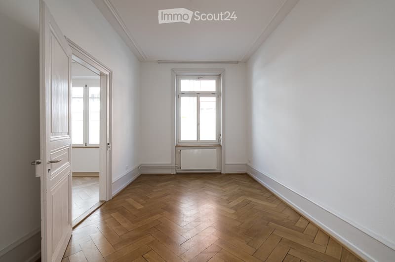 Empty room with light-colored walls, wooden herringbone flooring, and a single window with white frame. A door leads to another room with similar flooring.