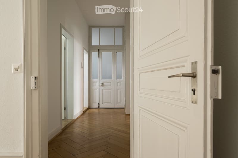 A clean, well-lit hallway with wooden flooring, white walls, and several doors including a mirrored door and a double door at the end.