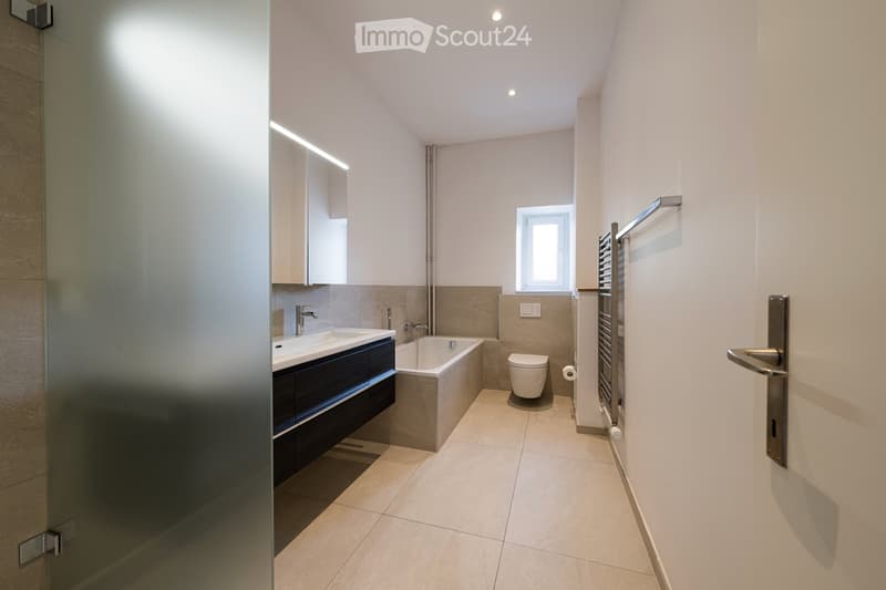 Modern bathroom with a double sink, bathtub, toilet, and a large mirror. The space features light-colored tiles and a window for natural light.