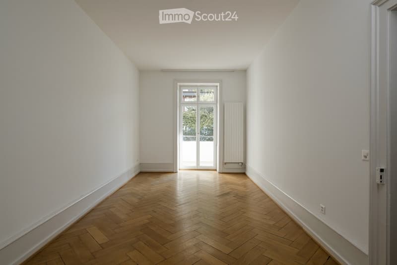 Empty room with wooden herringbone flooring, white walls, and a large window with a view of greenery outside. A radiator is visible on one wall.