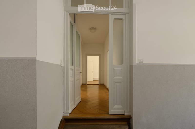A narrow hallway with wooden flooring, white walls, and a staircase leading upwards. The hallway features double doors with glass panels on either side.