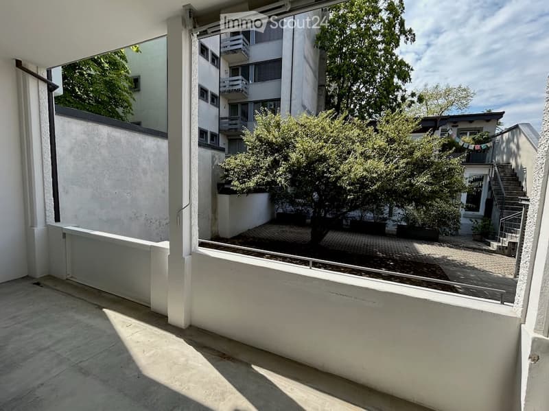 Balcony with modern glass railing, overlooking a tree-lined courtyard with buildings and greenery.