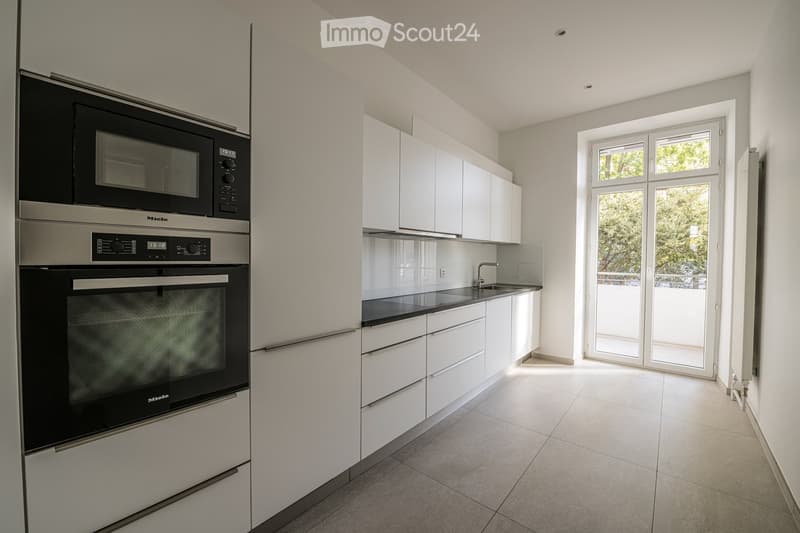 Modern kitchen with white cabinets, black countertops, stainless steel appliances, and a large window offering a view of greenery.