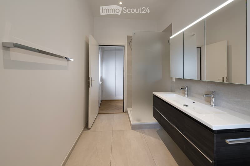 Modern bathroom with a double sink, sleek dark cabinetry, large mirror with LED lighting, and a visible hallway leading to another room.