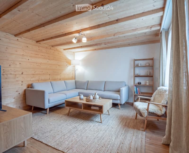 Modern living room with exposed wooden beams, light grey sectional sofa, wooden coffee table, and a cozy armchair with a fur throw. The room features wooden walls, a large window with white curtains, and a wooden shelf with decorative items.