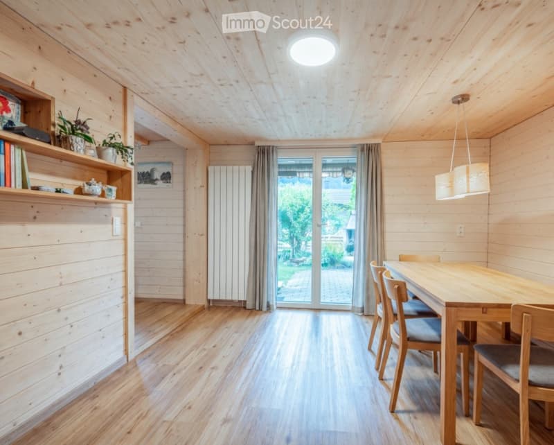 Bright wooden dining room with a long wooden dining table, chairs, a shelf with books and plants, and a large window offering a view of the garden.