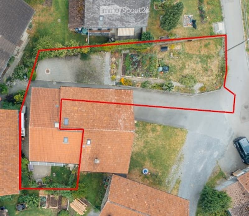 Aerial view of a residential building with a tiled roof, surrounded by a garden area and a driveway. The property is outlined in red, showing its boundaries.