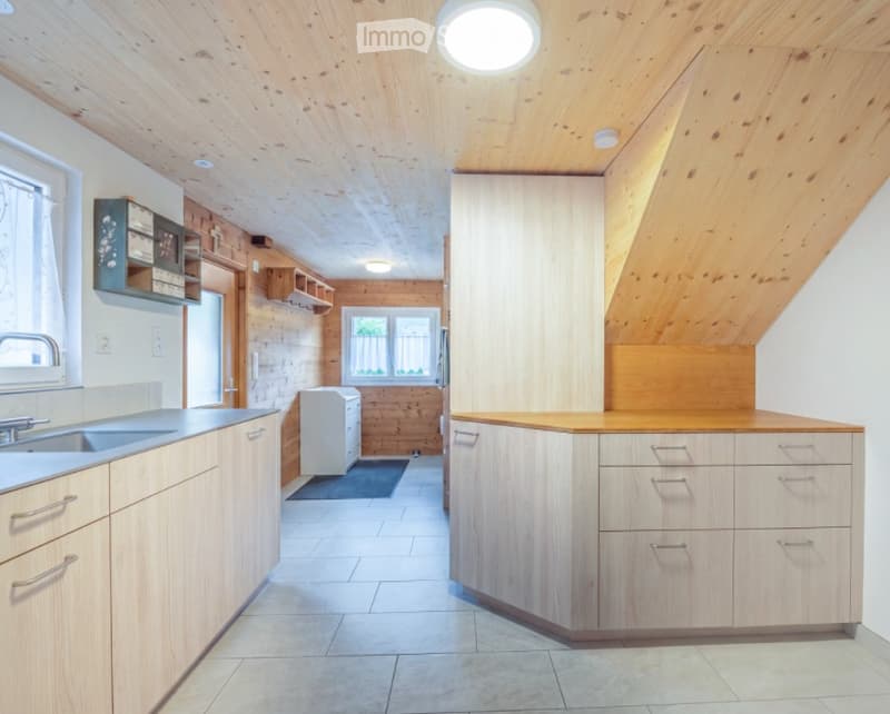 Bright, modern kitchen with wooden cabinets, stainless steel sink, and a small refrigerator tucked into a corner. The space features exposed wooden beams on the ceiling and a mix of wooden and tiled flooring.