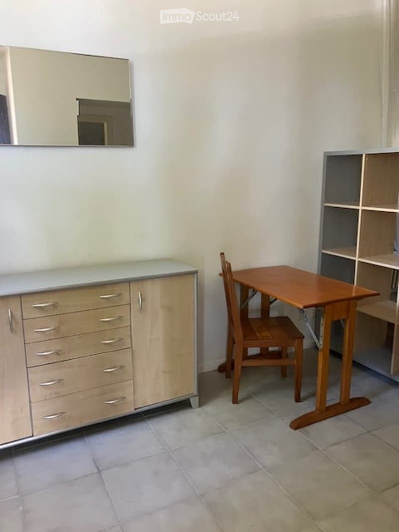 A small, furnished room with a light-colored cabinet, a wooden chair, a small table, and a shelf unit.