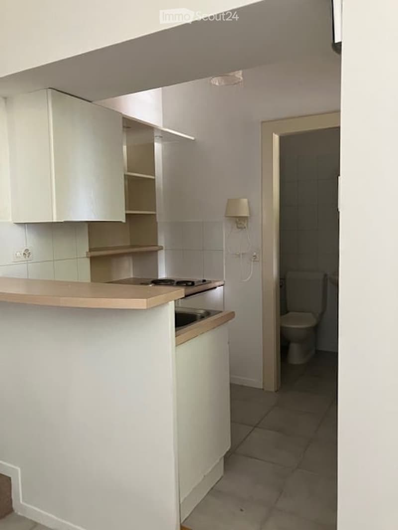 Modern kitchen with white cabinets, a countertop, and a sink. A small bathroom is visible through an open doorway, featuring a toilet and tiled walls.