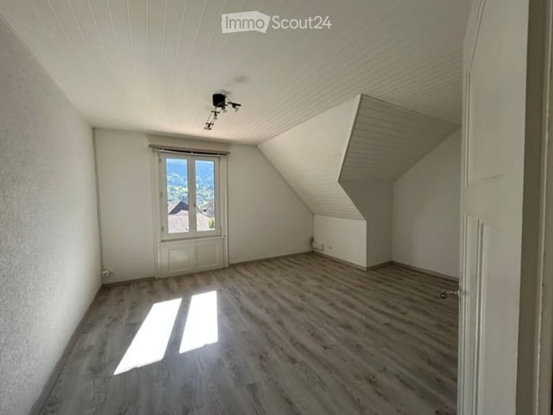 Empty attic room with wooden flooring, white walls, skylight window offering a view, and exposed wooden beams on the ceiling.