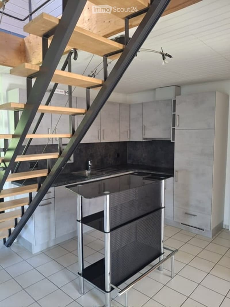 Modern kitchen with grey cabinets, a black countertop, a sink, and a bar stool. A wooden staircase leads to an upper level.