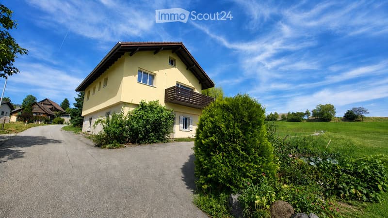 Two-story yellow house with a brown roof, multiple windows, and a balcony on the upper floor. The house is surrounded by greenery and is situated on a paved driveway.