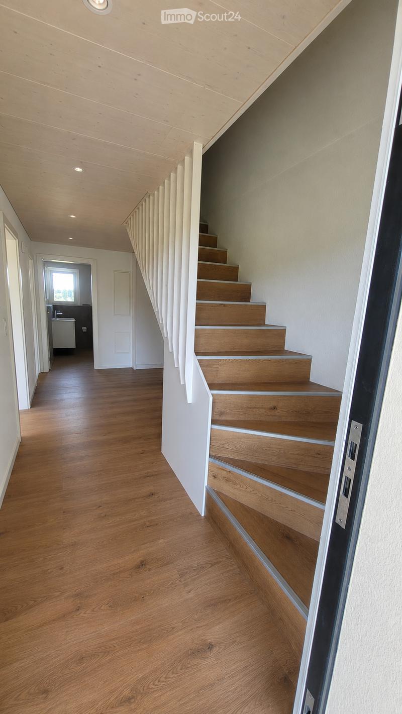 Spacious hallway with wooden flooring, white walls, and a wooden staircase leading upwards. The ceiling is made of light wood with recessed lighting.