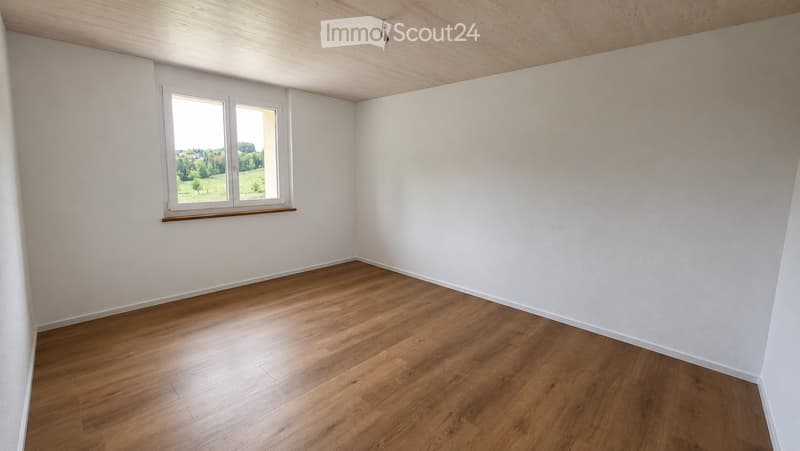 Empty room with wooden flooring, white walls, and a large window with a view of greenery outside.