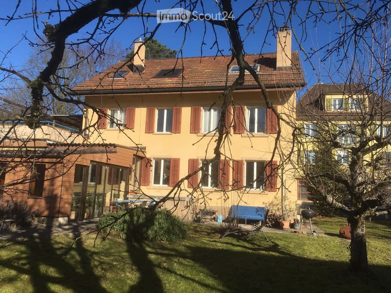 Two-story house with yellow facade, red shutters, and a tiled roof. The house features a well-maintained garden with a blue bench, a barbecue, and various plants.