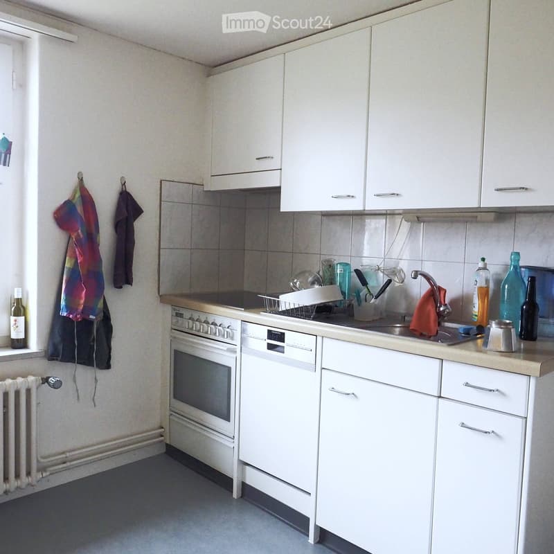 A small, modern kitchen with white cabinets, a built-in oven, a dishwasher, and a sink. The countertop has various items including cups, bottles, and a dish rack.