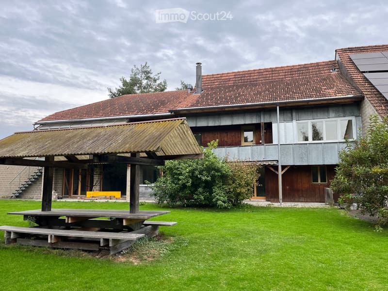 Two-story house with red-tiled roof, wooden and concrete exterior, and a covered outdoor seating area with a picnic table and benches.