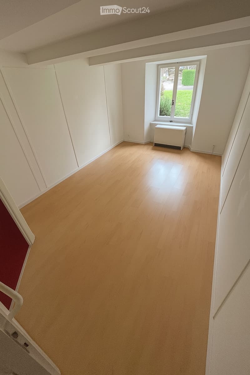 Empty room with wooden floor, white walls, a small white cabinet under the window, and a window with a small glass pane opening to the outside