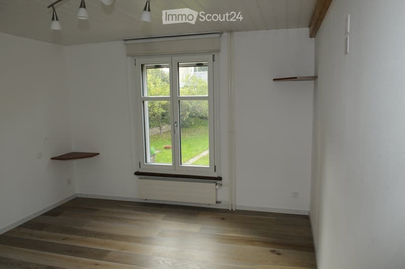 Empty room, white walls, wooden floors, window with wooden frame, shelf, radiator, view of garden outside