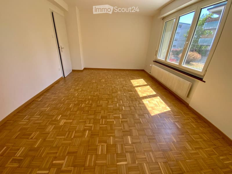 Empty room with wooden herringbone flooring, white walls, large windows, and a radiator. No furniture or equipment present.