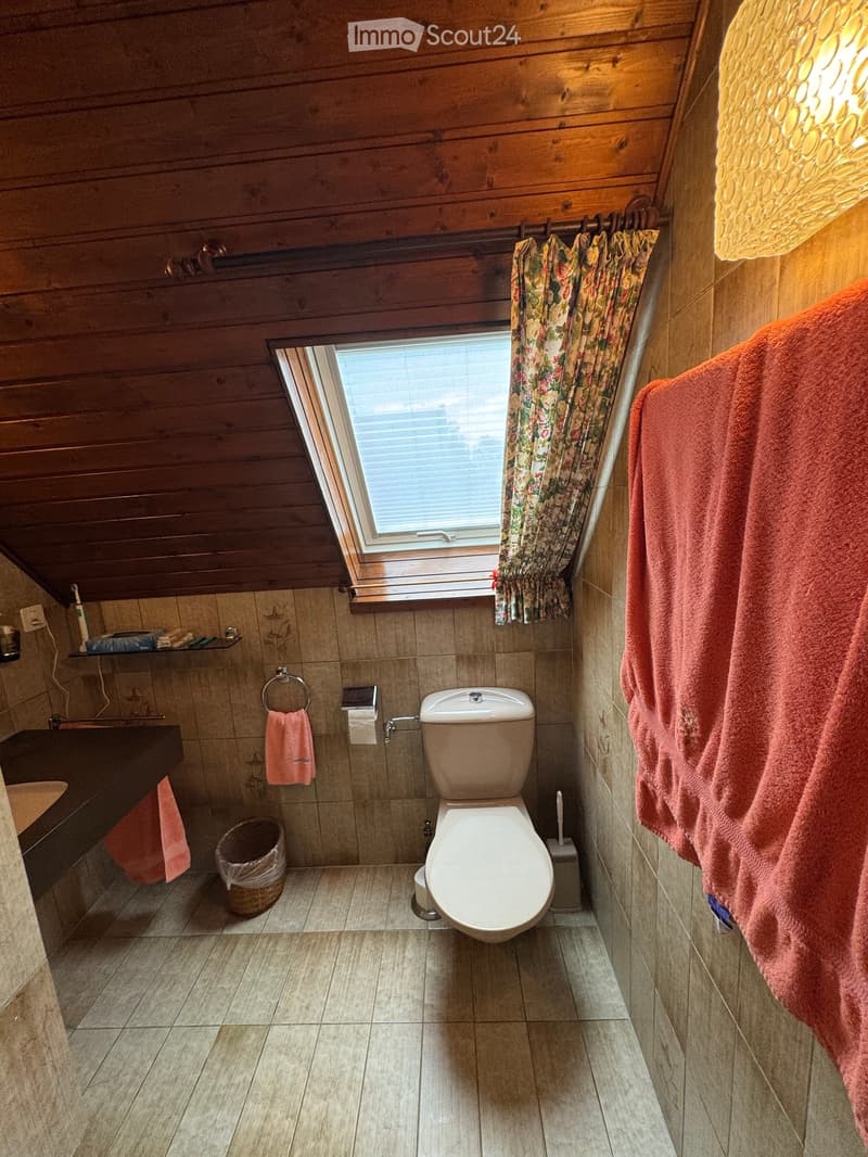 Bathroom with wooden ceiling, tiled walls, toilet, sink, and window with floral curtains. Towels and a small basket are visible.
