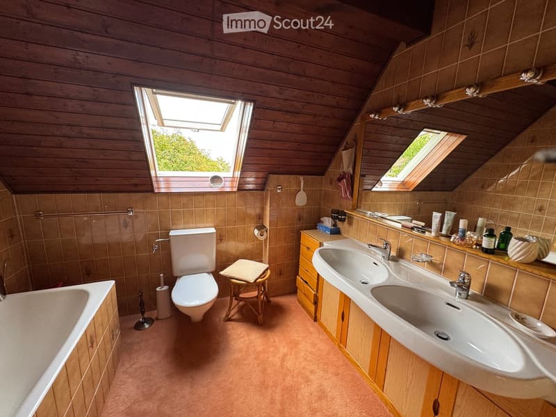 Bathroom with wooden ceiling, tiled walls, double sink, toilet, bathtub, and a small wooden stool with a towel.