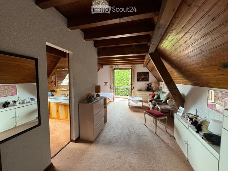 Spacious attic room with exposed wooden beams, carpeted floor, and large windows leading to a balcony. Features a sofa, coffee table, bed, cabinets, and decorative items.