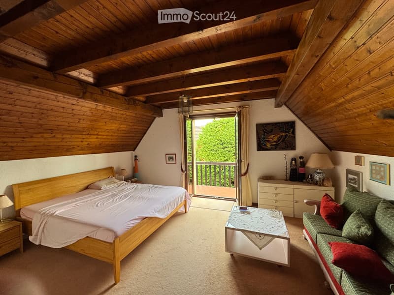 Cozy attic bedroom with wooden beams, a large bed, a sofa, a coffee table, and a balcony overlooking greenery.