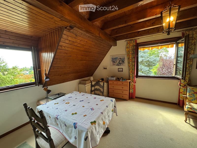 Cozy attic room with wooden beams, large windows, a table with a butterfly-patterned tablecloth, chairs, a lamp, and a chest of drawers.