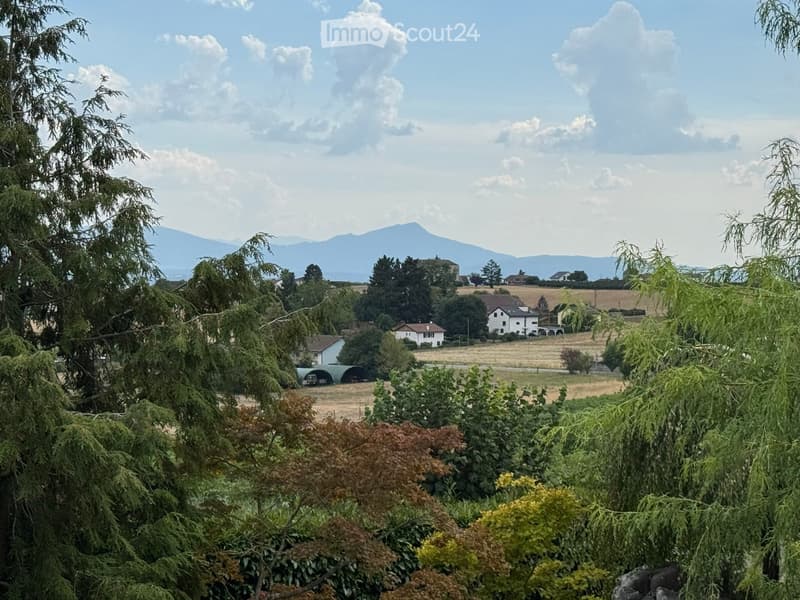 A rural landscape with scattered houses, trees, and a distant mountain range under a partly cloudy sky.