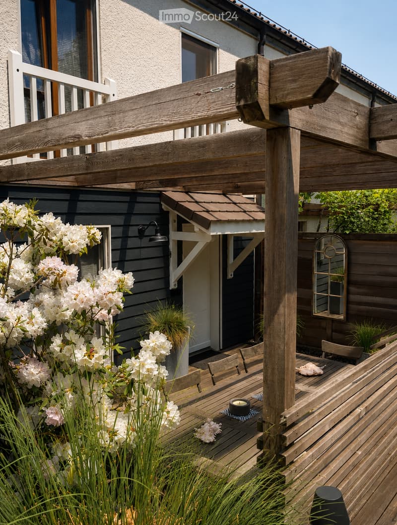 Wooden patio with a table, chairs, and a small candle, surrounded by white flowering shrubs and greenery, with a wooden pergola and a modern house in the background.