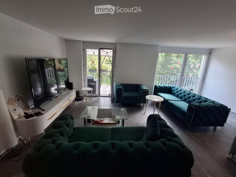Modern living room with teal sofas, glass coffee table, TV stand, side tables, and a lamp. Large windows and sliding glass doors lead to a balcony.