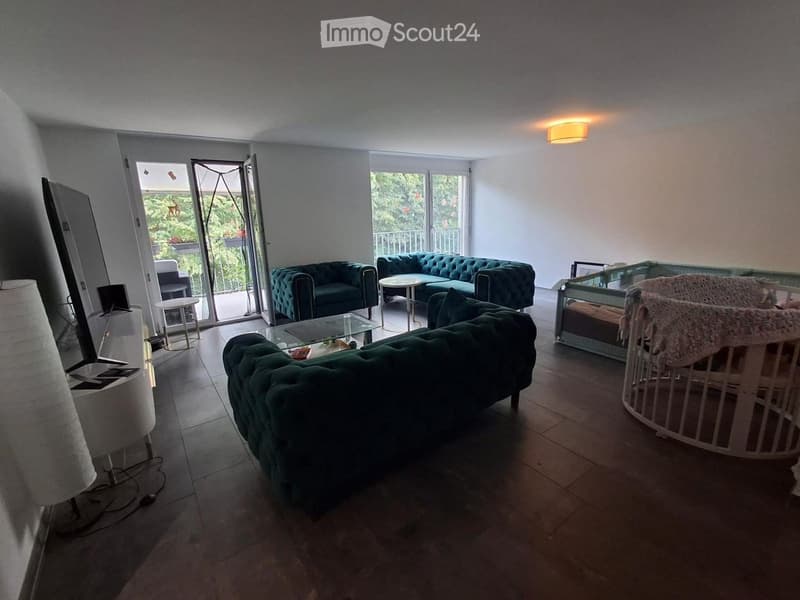 Modern living room with dark green sofas, glass coffee table, small round table, and a baby crib. Large sliding glass doors open to a balcony with outdoor seating.