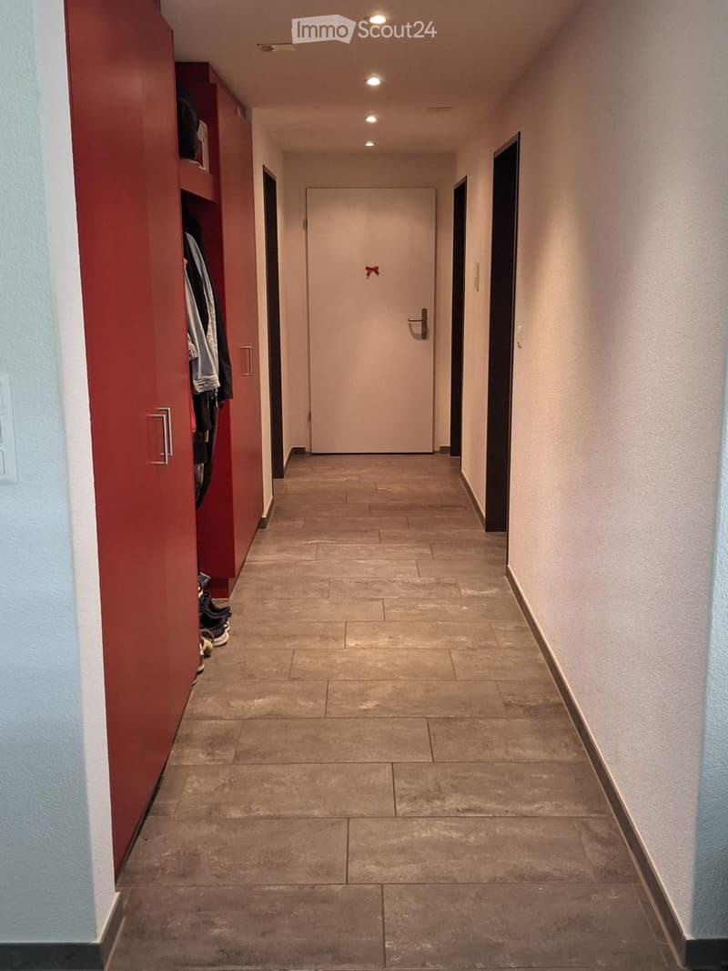 Clean hallway with tiled floor, beige walls, and a closed door at the end. A red wardrobe with hanging clothes is visible on the left side.