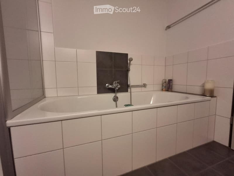 Bathtub with a modern faucet, a mirror, and a handrail for safety, with tiled walls and a dark floor.