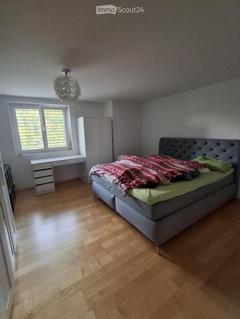 Spacious bedroom with wooden flooring, a large bed with a patterned bedspread and pillows, a modern desk with a chair, a wardrobe, and a decorative light fixture.
