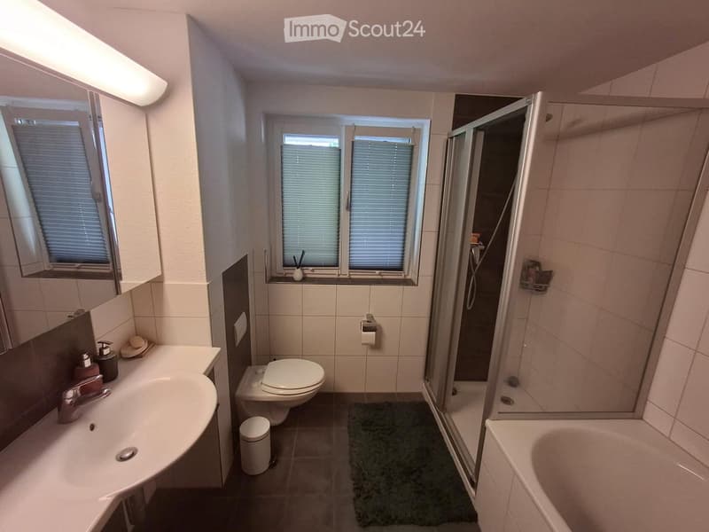 Modern bathroom with a sink, toilet, bathtub, and shower. Features include a large mirror, window with blinds, and a small trash bin.