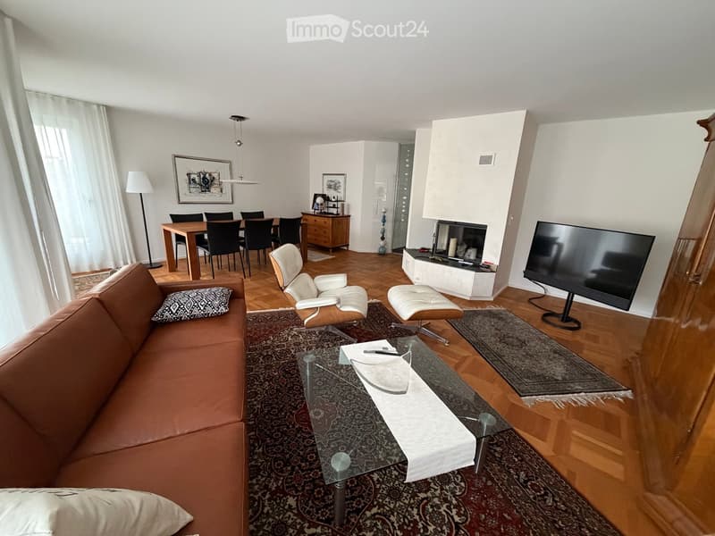 Modern living room with a brown leather sofa, glass coffee table, white armchair, white stool, flat-screen TV, dining area with a wooden table and chairs, and a wooden cabinet.