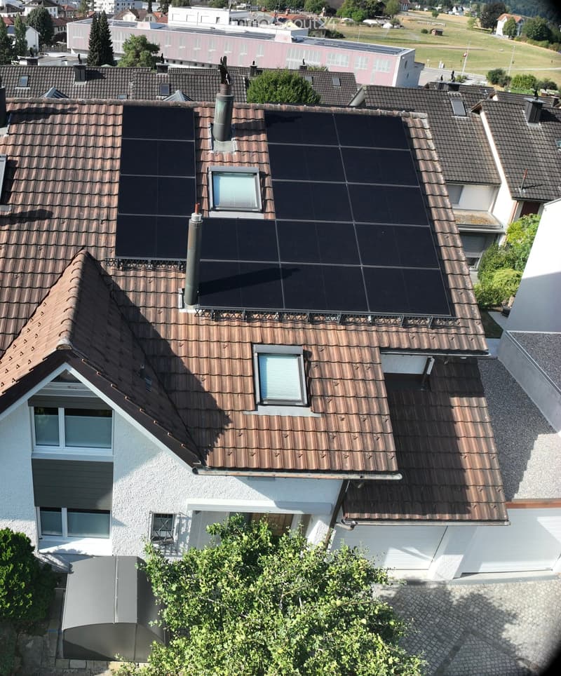 A multi-story residential building with a brown tiled roof, solar panels, chimneys, and multiple windows. The surrounding area includes other buildings, greenery, and open fields.